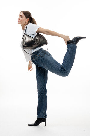 Woman in jeans, white t-shirt, and heeled boots balances on one leg, holding her other ankle behind her. She carries a sleek black INA KENT shoulder bag and stands against a plain white background.