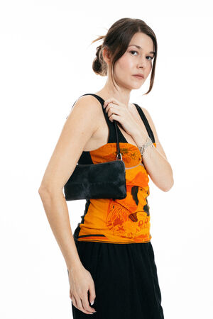 Woman with brown hair in an orange tank top and black skirt holds an INA KENT black handbag over her shoulder, posing against a white background.