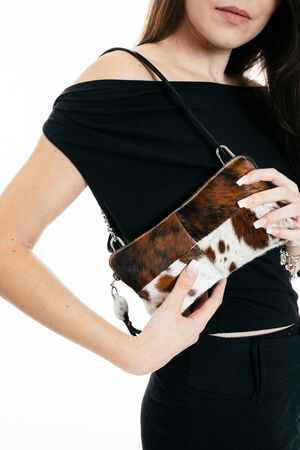 Woman in a black off-shoulder top holds an INA KENT small brown and white animal print purse with a thin black strap against a white background.