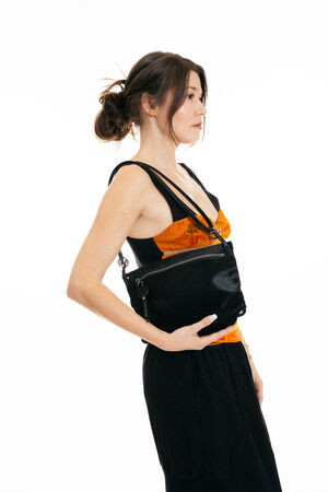 Woman in an orange and black top and black skirt stands sideways, holding an INA KENT black shoulder bag against a plain white background.