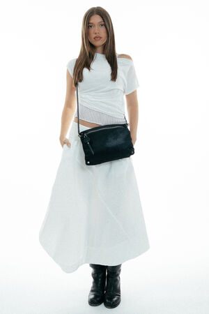 Woman wearing a white short-sleeve top, long white skirt, black boots, and carrying an INA KENT black crossbody bag, standing against a white background.