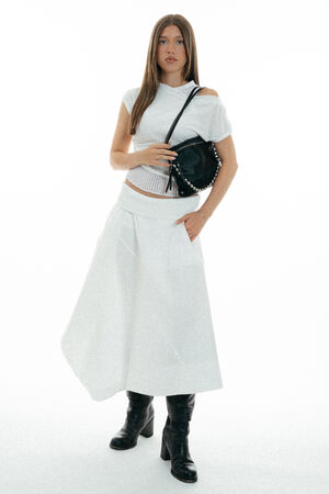 Woman standing against a white background wearing a white short-sleeve top, long skirt, black boots, and holding an INA KENT black handbag with a shoulder strap.