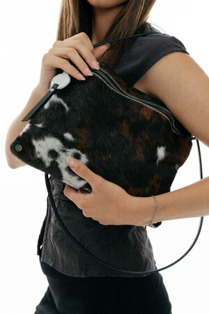 A person holds an INA KENT brown and white cowhide-patterned handbag with a zipper and black strap, wearing a short-sleeved dark top.