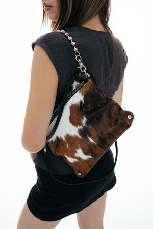 A person holds an INA KENT brown and white cowhide-patterned handbag with a zipper and black strap, wearing a short-sleeved dark top.