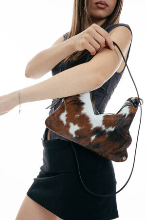 A person holds an INA KENT brown and white cowhide-patterned handbag with a zipper and black strap, wearing a short-sleeved dark top.