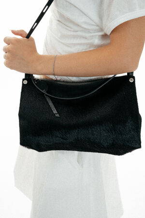 Person in a white outfit holding an INA KENT black textured shoulder bag with a zipper and thin strap, shown from the waist up against a white background.