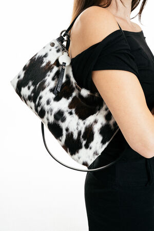 A woman in a black outfit carries an INA KENT black and white cow-print handbag over her shoulder against a white background.