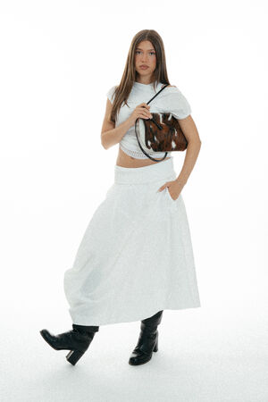 Woman standing against a white background, wearing a white top, long skirt, black boots, and holding an INA KENT brown patterned handbag.
