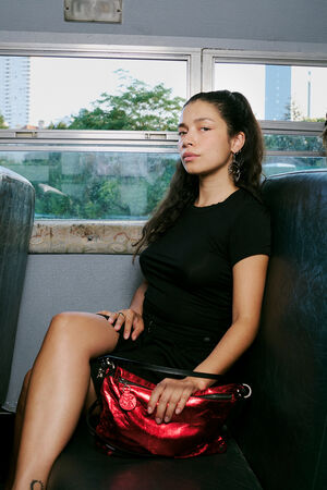 Three women sit separately on a bus, each appearing lost in thought. The woman in the center looks directly ahead, holding a shiny red INA KENT bag on her lap. Trees and buildings are visible through the windows.