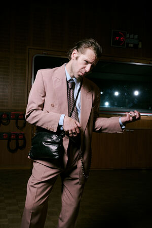Man in a pink suit pretends to play an air guitar with an INA KENT black handbag in a dimly lit room with soundproofing panels and clocks on the wall.