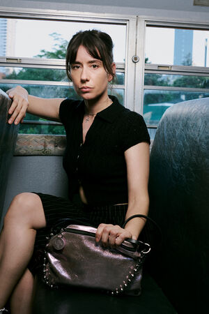 Three women sit separately on a bus; the woman in the center seat, her INA KENT handbag resting on her lap, looks directly at the camera, while the other two face away, each holding a handbag.