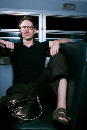 Two people sit on a bus; one sits casually with legs apart and arms on the seat, while the other sits upright wearing sunglasses. Both wear dark clothing, exuding an understated style that could easily be complemented by an INA KENT bag.