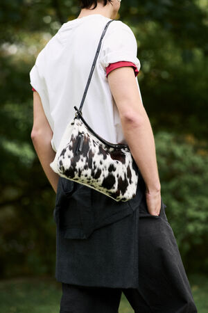 Person wearing a white t-shirt and dark pants carries an INA KENT black and white cow-print shoulder bag outdoors, with one hand in their pocket.