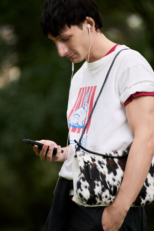 Person wearing headphones and a graphic T-shirt looks at their phone while holding an INA KENT black and white spotted bag, standing outdoors.