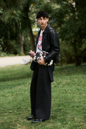 A person stands on grass holding a phone and an INA KENT black-and-white bag, wearing a black leather jacket, wide black pants, and black shoes, with trees in the background.