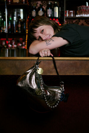 A person with short hair leans on a bar counter, smiling, with an INA KENT metallic handbag hanging from their arm. Bottles and glasses are visible in the background.