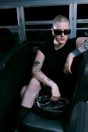 Person with short hair, sunglasses, and tattoos sits on a bus seat, holding an INA KENT bag. Two other people sit nearby, partially visible. The bus interior is dimly lit.