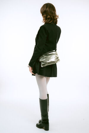 A person seen from behind, wearing a black blazer, a pleated skirt, white tights, and black boots, carrying a metallic silver crossbody bag.
