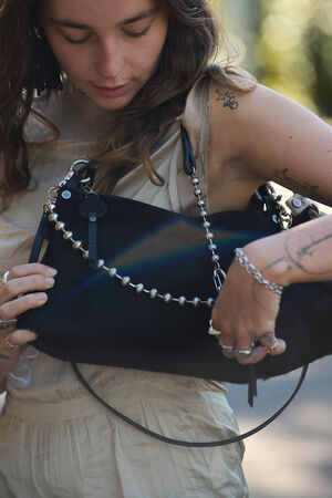 A woman in a sleeveless beige outfit holds a black bag with silver chain details, looking down as sunlight creates a rainbow on the bag.