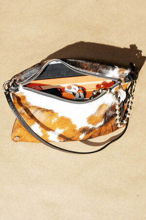 A cowhide leather handbag with a black strap, metal bead keychain, and visible personal items inside, placed on a light tan surface.