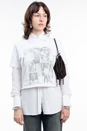 A person wears a white graphic T-shirt layered over a white button-down shirt, accessorized with a small dark INA KENT shoulder bag and silver jewelry.