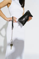 A person in a sleeveless white dress holds an INA KENT black clutch in one hand, with keys attached to a keychain clipped to their waist.